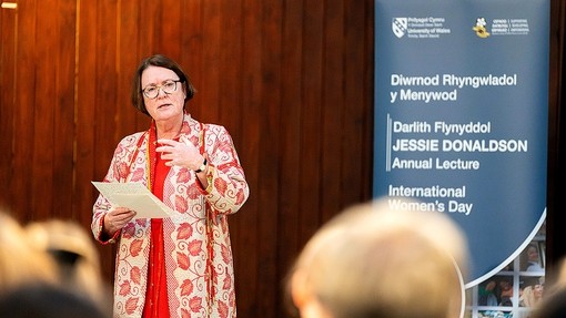 Yn gwisgo cot hir gyda phatrwm o ddail coch, gydag un llaw i fyny i bwysleisio, mae’r Athro Elwen Evans yn traddodi darlith ar thema ysbrydoli cynhwysiant.